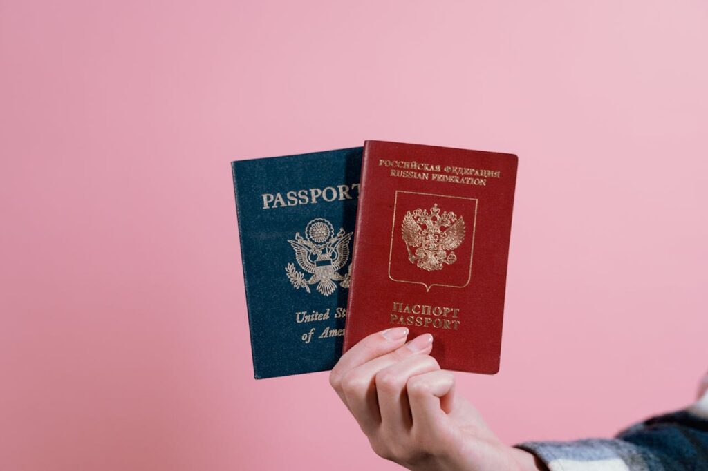 Close-up of a hand holding American and Russian passports on a pink background.